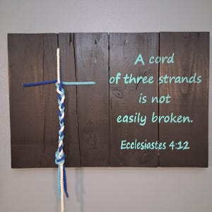 May include: Wooden wall art featuring a dark stained wood background with a cross design made of rope. The cross is made of blue, white, and light blue rope. Text in teal reads "A cord of three strands is not easily broken. Ecclesiastes 4:12."