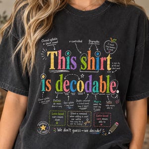 May include: A charcoal gray short-sleeve t-shirt featuring a colorful educational graphic with the text This shirt is decodable. The design includes various phonics rules, arrows, and labels such as Closed syllable, Digraphs, Prefixes, and Suffix.
