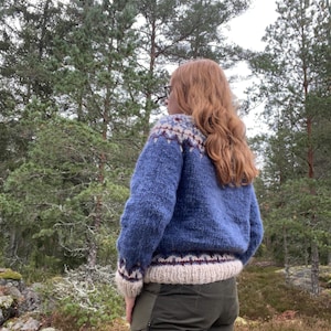Könnte beinhalten: Ein blauer Strickpullover mit gemustertem Passe und Bündchen. Der Pullover hat ein cremefarbenes Band an Taille und Bündchen mit burgunderfarbenen Akzenten. Die Person trägt eine olivgrüne Hose und steht in einem Wald.