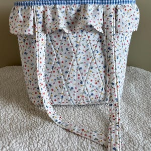 May include: A quilted tote bag with a white background and a floral pattern in red, blue, and yellow. The bag has a blue gingham trim and matching straps. The bag is approximately 12 inches tall and 10 inches wide.