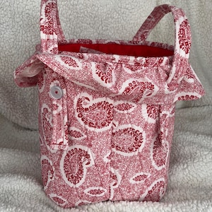 May include: A handmade fabric tote bag with a red and white paisley pattern. The bag has a red interior, two handles, and a button closure. The bag is made of quilted fabric.
