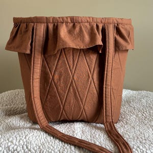 May include: A brown tote bag with a ruffled top and diamond-patterned stitching. The bag has two long straps and is made of a soft, textured fabric. The bag is sitting on a white, fluffy surface.