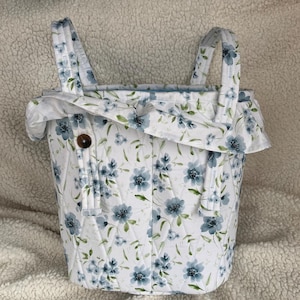 May include: A white quilted fabric basket with blue floral pattern and two handles. The basket has a ruffled top and a wooden button closure. The floral design includes blue flowers and green leaves.