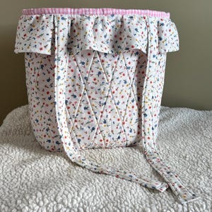 May include: A white fabric basket with a floral pattern of red, blue, and yellow flowers. The basket has a ruffled edge with a pink and white gingham trim and long fabric straps. The basket is quilted with a diamond pattern.