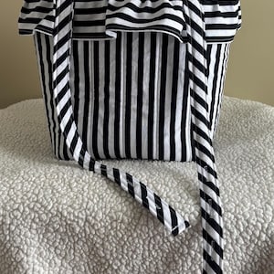 May include: A black and white striped tote bag with a ruffled top edge and matching straps. The bag has vertical stripes and is sitting on a white, textured surface. The straps are diagonal stripes.