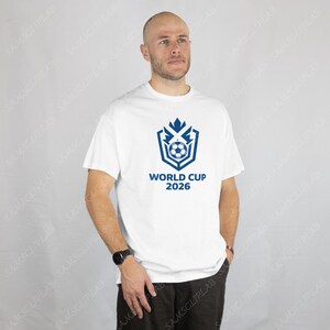 Puede incluir: Camiseta blanca con un gr&aacute;fico azul que representa un bal&oacute;n de f&uacute;tbol y el texto "WORLD CUP 2026". El dise&ntilde;o est&aacute; centrado en la camiseta. La camiseta est&aacute; hecha de un material suave.