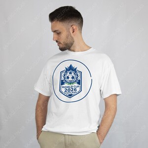 Puede incluir: Camiseta blanca con un dise&ntilde;o gr&aacute;fico azul y blanco para la Copa del Mundo 2026 en Escocia. El dise&ntilde;o incluye un bal&oacute;n de f&uacute;tbol, hojas de arce y el texto "WORLD CUP 2026 SCOTLAND" dentro de un c&iacute;rculo.