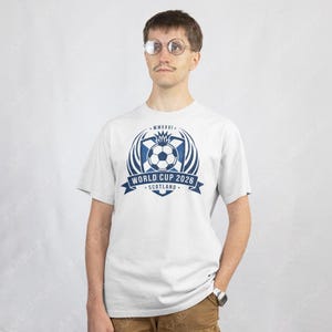 Puede incluir: Camiseta blanca con un dise&ntilde;o gr&aacute;fico azul y blanco para la Copa del Mundo 2026 en Escocia. El dise&ntilde;o incluye un bal&oacute;n de f&uacute;tbol, una pancarta con el texto "World Cup 2026 Scotland" y el n&uacute;mero romano "MMXXVI".