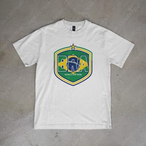 Puede incluir: Camiseta blanca con un diseño en forma de escudo en verde, amarillo y azul, que muestra las letras "BRA" y el texto "WORLD CUP 2026". El diseño incluye una estrella.
