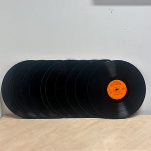 Pode incluir: Uma coleção de discos de vinil pretos, dispostos em fila. Um disco tem um rótulo laranja com texto e um logotipo. Os discos estão sobre uma superfície clara, com uma parede branca ao fundo.