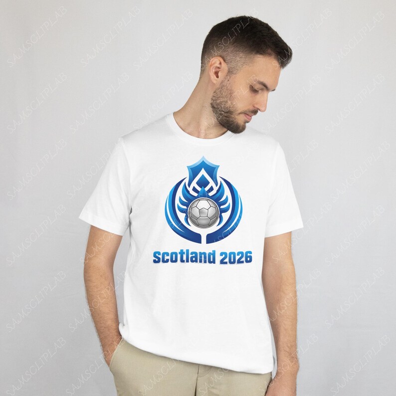 Puede incluir: Camiseta blanca con un dise&ntilde;o de bal&oacute;n de f&uacute;tbol azul y plateado y el texto "Scotland 2026". El gr&aacute;fico incluye un escudo y elementos en forma de alas. La camiseta la lleva una persona.