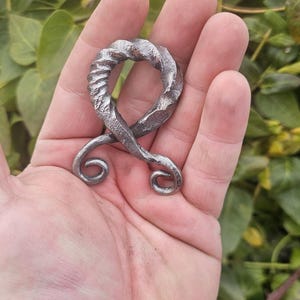 May include: A hand-forged, silver-colored metal pendant. The pendant features a twisted, textured top section and two curled bottom sections. The object is held in a person's hand against a backdrop of green foliage.