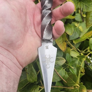 May include: Hand-forged metal dagger with a twisted handle and a sharp, pointed blade. The blade features etched symbols and a decorative design. The dagger is a dark, metallic color, and the handle has a textured appearance.