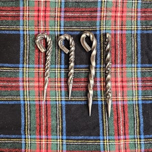 May include: Four hand-forged metal skewers with a twisted design and pointed ends. The skewers are silver and rest on a black, red, green, and blue plaid fabric.