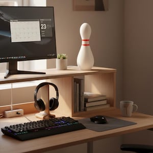 Puede incluir: Una moderna configuración de oficina en casa con un monitor de computadora que muestra un calendario, una lámpara blanca con forma de bolos con rayas rojas, un teclado, auriculares y un ratón. El escritorio es de madera clara.