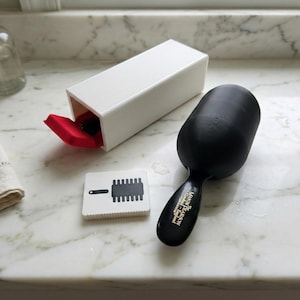 May include: A black Mason Pearson hairbrush with a long handle and a white storage box. The box is open, revealing a red interior. A small white card with a brush illustration is also present.