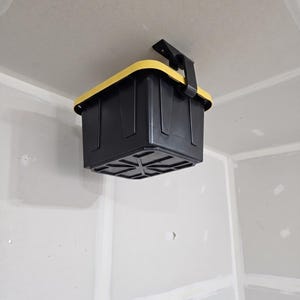May include: A black storage bin with a yellow rim is mounted on a ceiling bracket. The bin is rectangular and appears to be made of plastic. The bracket is also black and is attached to the ceiling. The bin is hanging upside down.