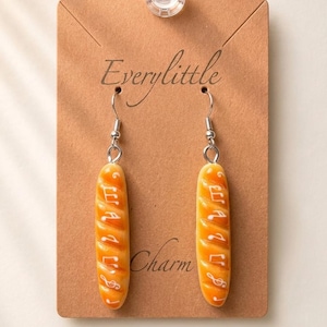 May include: A pair of dangle earrings featuring miniature baguette-shaped charms in a golden-brown color. Each baguette is decorated with white musical notes and symbols. The earrings are attached to a brown display card labeled Everylittle Charm.
