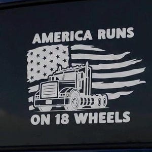 May include: A white vinyl decal applied to a dark surface featuring a semi-truck silhouette positioned in front of a distressed American flag. The text AMERICA RUNS is printed above the truck and ON 18 WHEELS is printed below the truck graphic.