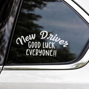 Op de afbeelding: Witte vinyl sticker voor een autoruit met de tekst "New Driver GOOD LUCK EVERYONE!!" in een mix van cursieve en schreefloze lettertypen. De sticker is aangebracht op een licht getinte autoruit.