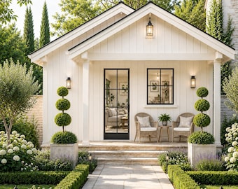 May include: A white cottage with vertical siding, a black-framed glass door, and a matching window. Two wicker chairs with cushions and a small table sit on the porch. Two topiary trees in square planters flank the entrance steps.