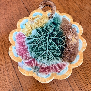 May include: A ceramic dish shaped like a cluster of leaves with a central handle. The surface features a textured leaf vein pattern in shades of green, brown, and pink, surrounded by a scalloped yellow border with light blue accents.