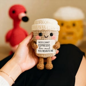 Op de afbeelding: Een gehaakte koffiekop met een lachend gezicht en een wit deksel. De beker heeft een klein bordje met de tekst "WORDS CANNOT ESPRESSO how much YOU MEAN TO ME". Een rode flamingo en een gele mok staan op de achtergrond.