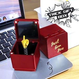 Könnte beinhalten: Eine rote Trick-Geschenkbox mit einer goldfarbenen Hand, die den Mittelfinger zeigt. Die Box ist geöffnet und zeigt die Hand auf einer Feder. Auf der geschlossenen Box steht in goldener Schrift "Just for You". Im Hintergrund ist ein Laptop zu sehen.
