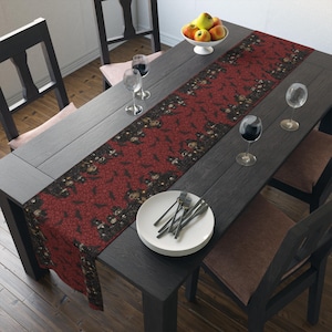 May include: A rectangular table runner featuring a dark red background with a repeating pattern of black bats and small, detailed illustrations of skulls and pumpkins. The runner is laid out on a dark wooden table with plates and glasses.