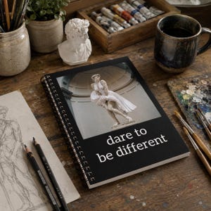 May include: A spiral-bound notebook with a black cover featuring a central image of a classical statue in a white dress and the white text "dare to be different" printed below the image. The notebook is positioned on a wooden surface.