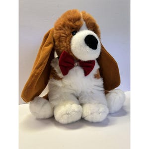 May include: A plush toy dog with brown and white fur, long floppy ears, and a red bow tie. The dog has a white muzzle and paws, with a black nose and eyes. The stuffed animal is sitting upright.