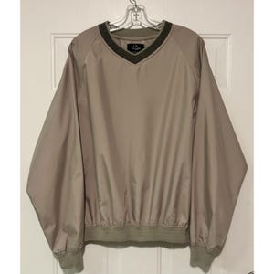 May include: Tan V-neck pullover windbreaker with a green collar and cuffs. The windbreaker has long sleeves and a drawstring hem. The brand logo is visible on the chest. This lightweight jacket is suitable for outdoor activities.