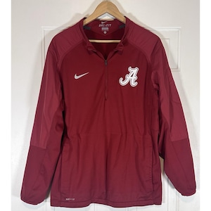 May include: A maroon Nike Dri-FIT long-sleeve pullover jacket featuring a quarter-zip neckline. The front displays a white Nike swoosh logo on the right chest and a white script letter A logo on the left chest. The garment is displayed on a hanger.