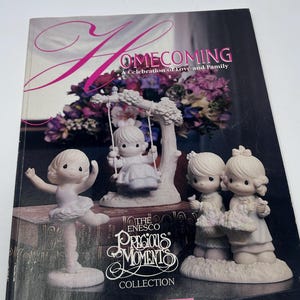 以下が含まれることがあります： 「Homecoming: A Celebration of Love and Family」というタイトルのソフトカバー本で、Enesco Precious Moments Collection Volume XIIIが掲載されています。表紙には、ダンサーやブランコに乗った子供たちなど、4体の磁器風のフィギュアが描かれています。