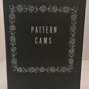 Puede incluir: Una caja negra con las palabras "PATTERN CAMS" en blanco, enmarcada por un delicado borde floral blanco. El diseño presenta una disposición cuadrada de rosas y hojas, creando una estética elegante.