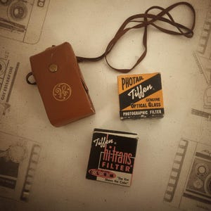 May include: A vintage camera case, brown leather with a gold emblem, sits with two Tiffen filter boxes. One box is black and orange, the other black and pink. A brown leather strap is also present.