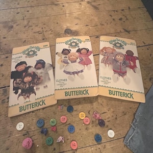 Könnte beinhalten: Drei Vintage-Butterick-Schnittmusterumschläge für Cabbage Patch Kids-Kleidung. Die Umschläge sind cremefarben mit Illustrationen der Puppen. Eine Vielzahl bunter Knöpfe sind unter den Mustern auf einer Holzoberfläche verstreut.