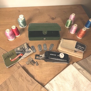 Op de afbeelding: Een verzameling vintage Singer naaibenodigdheden, waaronder spoelen garen in diverse kleuren, een groene opbergkoffer en een zwarte knoopsgatenmaker. Ook inbegrepen zijn instructieboekjes en metalen hulpstukken, alles gerangschikt op een houten oppervlak.