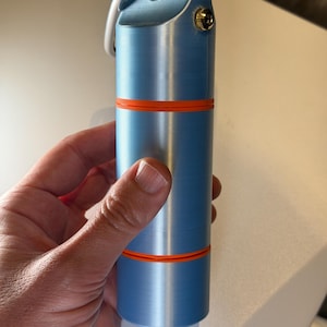 Puede incluir: Un objeto cilíndrico azul claro con bandas naranjas y un llavero plateado. El objeto tiene un pequeño botón dorado y una parte superior redondeada. El artículo se sostiene en una mano.