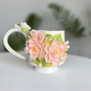 May include: White ceramic mug with a handle and a pedestal base, decorated with 3D sculpted pink flowers, yellow centres, and green leaves. The flowers are clustered on the front and handle, creating a decorative floral design. A unique and elegant teacup.
