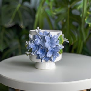 Può includere: Un vaso di ceramica bianco con base a piedistallo, adornato con fiori di ortensia blu e foglie verdi. La composizione floreale è centrata sul vaso, creando un accento decorativo. Il vaso è su un tavolo bianco.