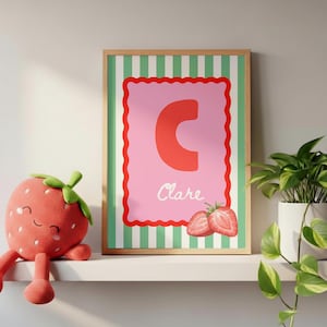 Op de afbeelding: Een ingelijste print met een rode letter "C" op een roze achtergrond en de naam "Clare". De lijst heeft een groen-witte gestreepte rand. Een aardbei knuffel en een potplant staan op een witte plank.