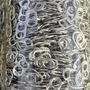 May include: A clear glass jar filled with numerous silver aluminum can pull tabs. The tabs are densely packed, creating a textured, metallic appearance. The jar's contents are illuminated, highlighting the shiny surfaces of the tabs.