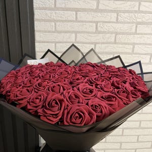 Könnte beinhalten: Ein großer Strauß tiefroter Rosen, eng arrangiert und in schwarzes Papier mit einer schwarzen Schleife gewickelt. Die Rosen stehen im Mittelpunkt, mit einer Backsteinmauer im Hintergrund.