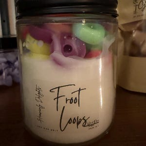 May include: A clear glass jar candle with a black lid, filled with white wax and colorful wax melts. The label reads "Froot Loops" and "Heavenly Delights". The wax melts are shaped like colorful cereal pieces.