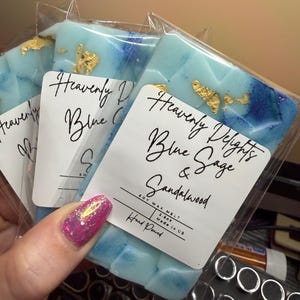 May include: Three rectangular soy wax melts in clear plastic packaging. The wax is light blue with dark blue swirls and gold leaf flakes. Each label reads Heavenly Delights Blue Sage & Sandalwood, Soy Wax Melt, 2.5oz, Hand Poured, Made in US.