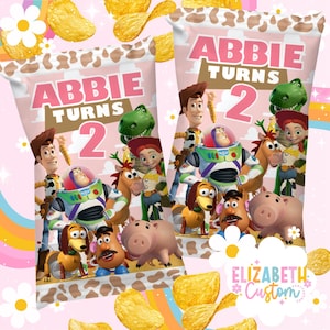 Puede incluir: Dos bolsas de patatas fritas rosas con el texto "Abbie Turns 2" y varios personajes de Toy Story. Las bolsas tienen un dise&ntilde;o de vaca en la parte superior y est&aacute;n rodeadas de patatas fritas y decoraciones florales.