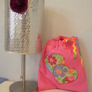 Puede incluir: Una pantalla de lámpara plateada con lentejuelas y una flor rosa en la parte superior, junto a una bolsa de tela rosa con dos aplicaciones de corazón de colores.