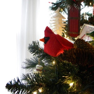 Puede incluir: Adorno navideño de un cardenal rojo brillante con pico y ojo negros, posado en una rama de árbol de Navidad. El árbol está decorado con luces, piñas y otros adornos festivos, creando una escena navideña.