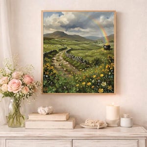 Könnte beinhalten: Ein gerahmtes Landschaftsgemälde zeigt eine lebendige irische Landschaft. Ein Regenbogen spannt sich über grüne Hügel, einen gewundenen Steinweg und einen Goldtopf. Gelbe Wildblumen und vierblättrige Kleeblätter säumen den Vordergrund. Das Kunstwerk wird auf einer weißen Oberfläche präsentiert.